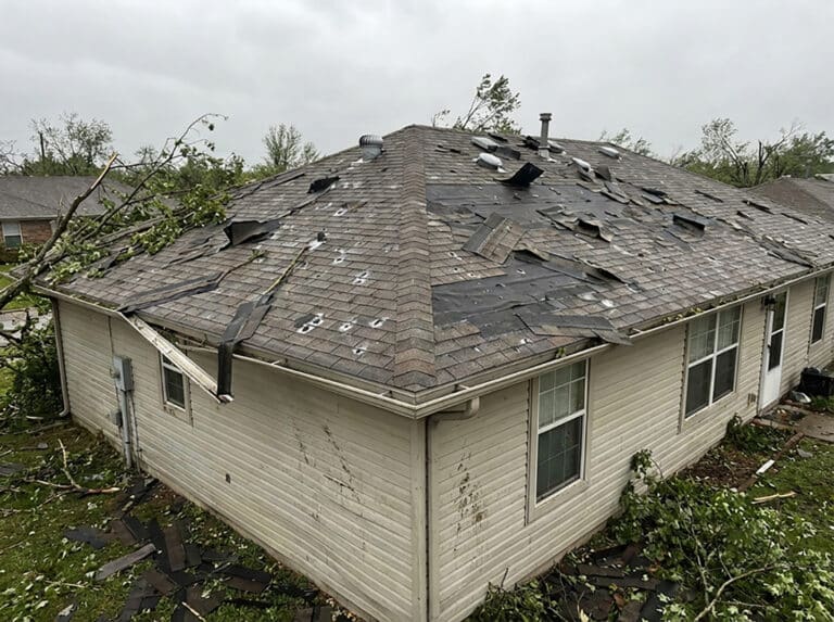 A home with severe storm roof damage showing torn shingles, exposed underlayment, scattered debris, and fallen branches across the roof, highlighting the need for storm damage roof repair.